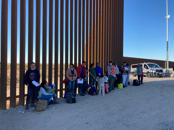 Familias y adultos que viajan solos esperan a lo largo del muro fronterizo con documentos a la mano para ser procesados por un agente de la Patrulla Fronteriza. El grupo de alrededor de 30 personas cruzaron la frontera de manera ilegal cerca de Somerton, Arizona, el 29 de noviembre de 2021.